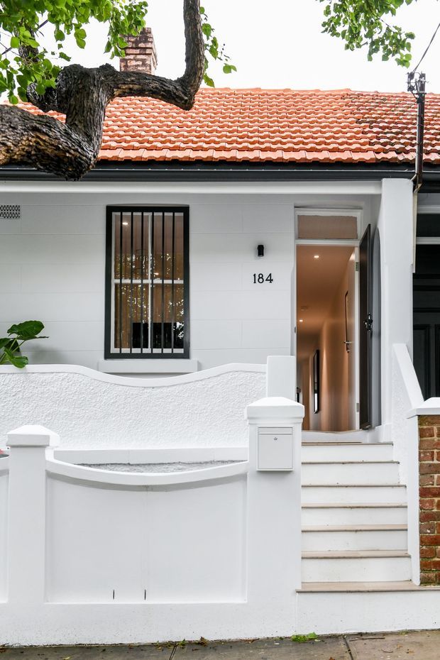 Street-facing view of a compact white rendered terrace or bungalow-style residence. The facade features a curved low boundary wall, a raised entry stair, black metal-barred window, open timber entry door, white mailbox, exposed brick side wall, and a terracotta tiled pitched roof partly shaded by a mature tree.