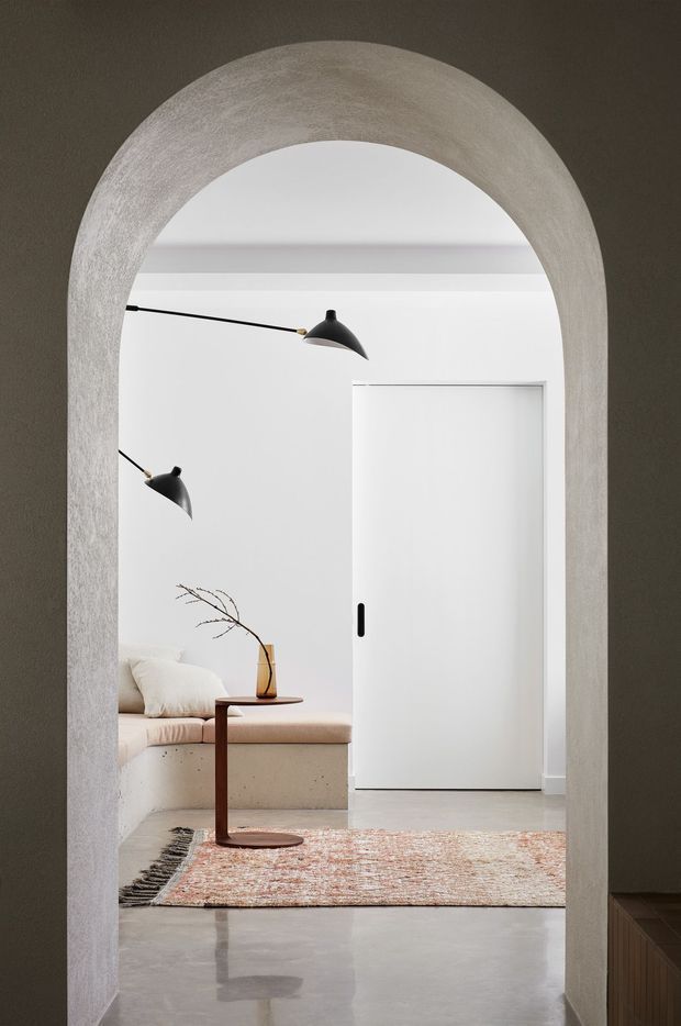 A softly lit minimalist interior viewed through a tall rounded plaster archway. The room beyond features white walls and ceiling, a concealed white sliding door, polished concrete flooring, a built-in upholstered bench or sofa, a slim dark wood side table, black wall-mounted lamps, and a muted patterned rug. The composition emphasizes the sculptural arch, restrained palette, and clean contemporary detailing.