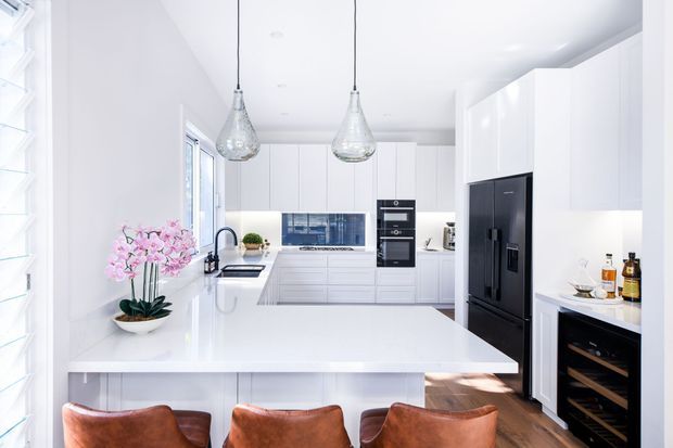 Bright, spacious contemporary white kitchen with a large waterfall-edge island, integrated white cabinetry, black appliances, a blue-toned glass or tiled splashback, pendant lighting, and warm timber flooring. Brown leather bar stools and a floral arrangement add contrast.