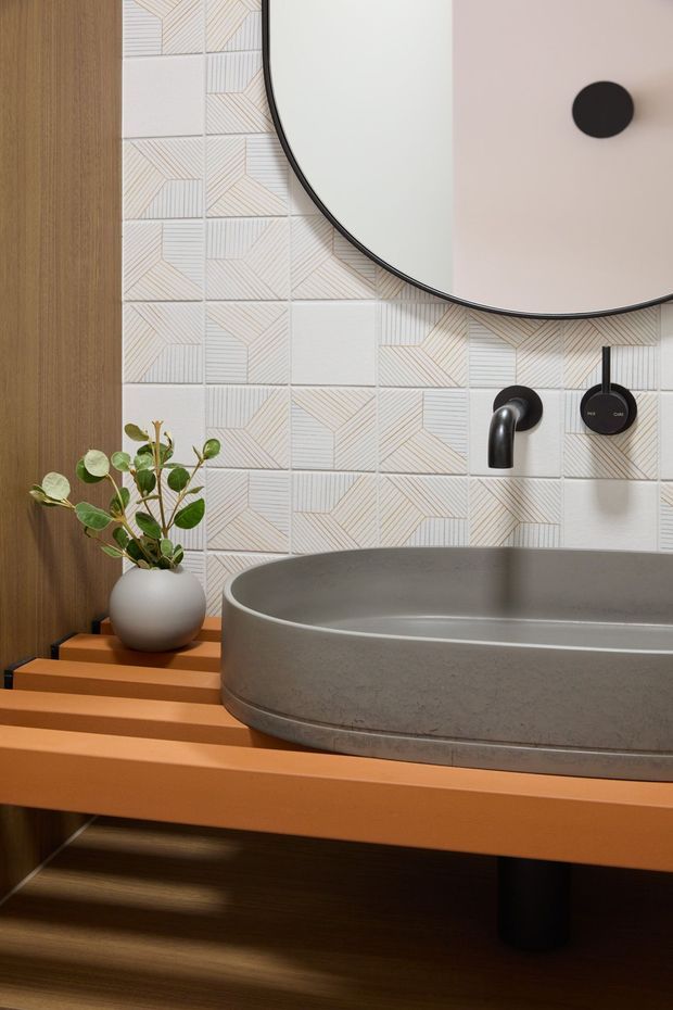Close-up view of a stylish bathroom vanity with a large round vessel basin in a warm grey concrete-like finish, wall-mounted dark metal tapware, and a round mirror. The vanity has a vivid terracotta-orange benchtop and timber cabinetry or shelving beneath. The backsplash features patterned geometric white tiles with fine linear detailing, while a small potted plant adds greenery. The composition is warm, refined, and contemporary with a strong material and colour contrast.
