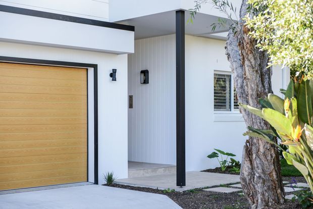 Contemporary residential entrance viewed from outside, featuring crisp white rendered walls, a flat-roofed projecting porch, black trim and supports, a warm timber-look garage door, vertical wall cladding at the entry, and landscaped planting around the walkway.