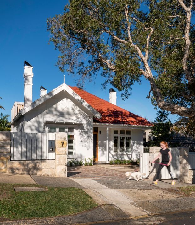Street-facing view of a renovated single-storey heritage-style home with a white rendered facade, steep gabled roof, prominent chimneys, red terracotta roof tiles, timber shutters and a low masonry boundary wall. A paved driveway and front garden sit beneath mature trees, with a pedestrian and dog passing in the foreground.