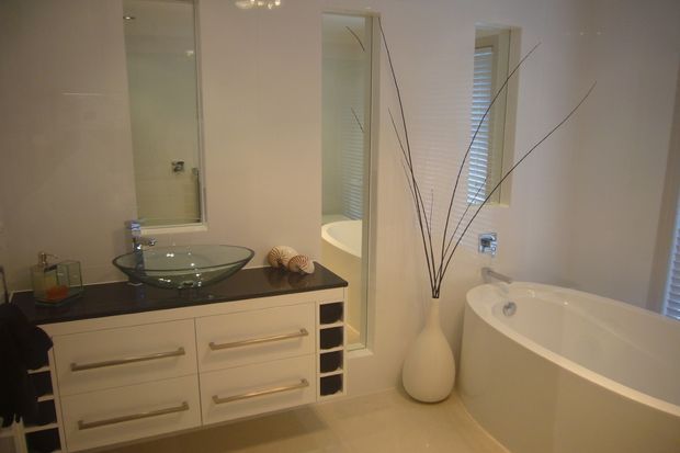 Bright contemporary bathroom featuring a freestanding white bathtub, glass vessel basin on a dark benchtop, mirrored wall cabinetry, white built-in vanity drawers, recessed shelving, and narrow windows with plantation shutters. Neutral beige and white finishes create a clean, refined atmosphere.