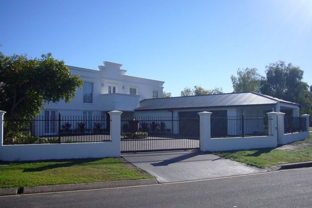 Street-facing view of a large white rendered residence with a low-pitched dark metal roof, projecting upper-level architectural elements, and a secure black metal fence with rendered masonry pillars. A paved driveway leads through a vehicle gate to the front of the property, with established trees and lawn surrounding the building.
