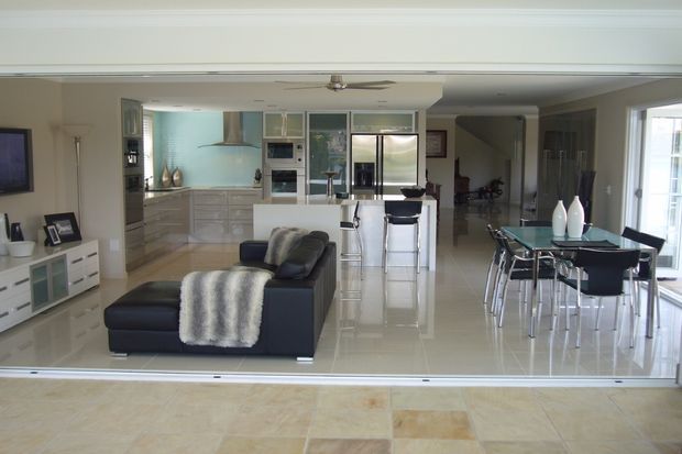 Open-plan contemporary home interior viewed from an outdoor terrace through wide sliding glass doors. The space combines a polished light-tile living area with a dark sectional sofa, a streamlined white kitchen with island seating, and a glass dining table with black chairs. Large glazing, pale finishes, and reflective surfaces create a bright, spacious feel.