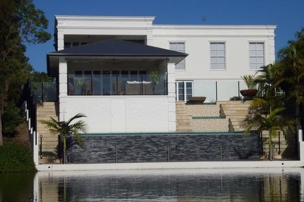 Front exterior view of a white two-storey residence beside a reflective body of water, featuring a flat roof with classical stepped parapet detailing, large rectangular windows, a covered upper-level terrace, broad tiled steps, dark stacked-stone boundary cladding, and tropical planting.