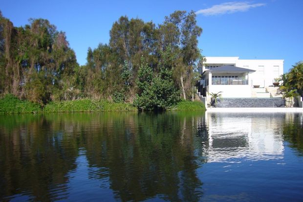 A modern white multi-level waterfront house viewed across a calm body of water, with broad glazing, a projecting flat-roofed upper section, a stone-clad lower wall, and landscaped vegetation surrounding the property. The building and trees are reflected in the water under a clear blue sky.