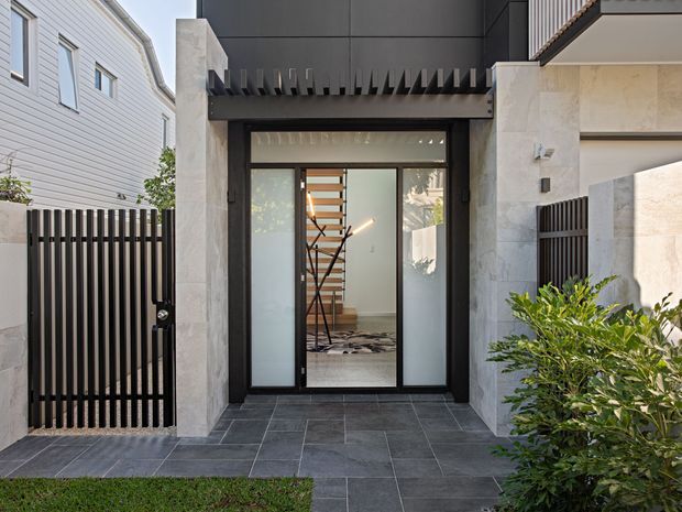 A contemporary residential front entry viewed from outside, featuring a dark framed glazed pivot or hinged door, pale stone-clad walls, charcoal tiled paving, vertical black metal fencing, and a sheltered entry canopy with projecting sunshade fins. Through the glass, a sculptural open staircase is visible inside.