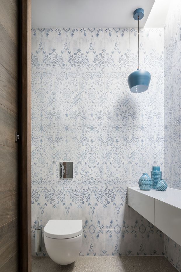 A compact contemporary powder room with a wall-hung white toilet, floating white vanity, patterned blue-and-white wall tiles, grey floor, dark timber door, and a blue pendant light. Decorative blue vessels sit on the vanity, while a mirrored flush plate and small toilet brush are visible.