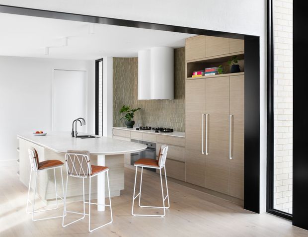 Bright contemporary kitchen with a compact white island and integrated cooktop, light timber full-height cabinetry, pale stone benchtops, and a textured vertical tile splashback. White bar stools with warm timber seats surround the island, while a large black-framed glazed opening adds strong contrast. The space is clean, open, and filled with natural light.