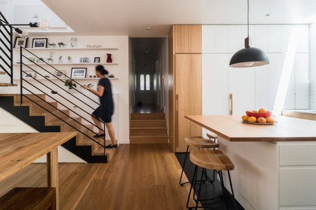 Bright open-plan interior centered on a contemporary kitchen and dining area, with a timber island and stools in the foreground, white handleless cabinetry, integrated vertical timber joinery, and a timber dining table. A black metal open-riser staircase with timber treads and a shelving wall occupies the left side, leading toward a darker hallway and entry beyond. The space uses clean lines, warm timber flooring and joinery, white surfaces, and black metal accents.