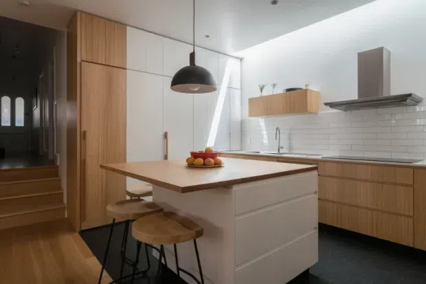 A bright contemporary kitchen with a central island, integrated white cabinetry, warm timber pantry and drawer fronts, and a white subway-tile splashback. The island has a timber benchtop with stools, while a dark pendant light, stainless-steel rangehood, timber flooring, and a dark floor section add contrast. A hallway and stairway are visible to the left.