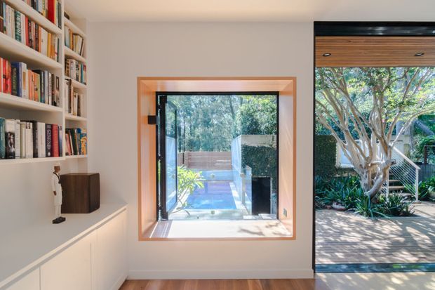 Bright contemporary interior with built-in white bookcases and lower cabinetry along the left wall, a compact dark wood speaker or cabinet, and warm timber flooring. A timber-framed window and large glazed opening provide views and access toward a landscaped courtyard with timber decking, mature trees, and a small pool or water feature. The space has clean built-in joinery and strong indoor-outdoor connection.