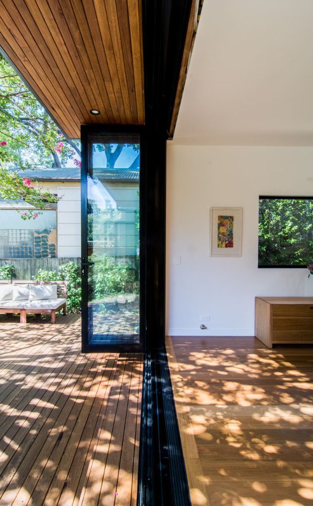 A bright indoor-outdoor living space divided by a large black-framed glazed opening. The interior has warm timber flooring, white walls, a timber ceiling soffit, and a simple timber media console. The adjoining timber deck extends into a leafy garden courtyard, with strong sunlight and dappled shadows throughout.