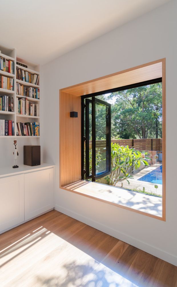 A bright, minimalist interior featuring a built-in white bookshelf and low cabinetry beside a large timber-lined window opening. The window has black-framed glazing and an open door, looking onto a timber deck, planted garden and a partially visible swimming pool. Warm timber flooring and window reveals contrast with pale walls, while strong sunlight creates patterned shadows across the floor.