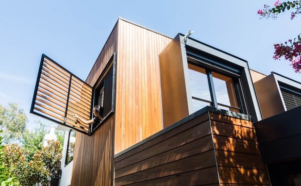Exterior architectural photograph of a contemporary multi-level building with vertically clad warm timber walls, darker horizontal timber cladding at the lower level, black-framed windows, and projecting operable louvre screens. The low-angle view emphasizes the angular facade and modern detailing against a clear sky.