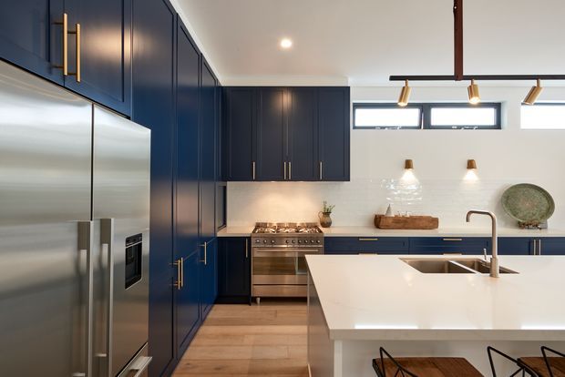 A bright contemporary kitchen with deep navy-blue floor-to-ceiling cabinetry, brushed metal appliances, a large white island with an integrated sink, pale timber flooring, and a white textured tile backsplash. Brass hardware, under-cabinet lighting, wall sconces, and a linear pendant add refined contrast.