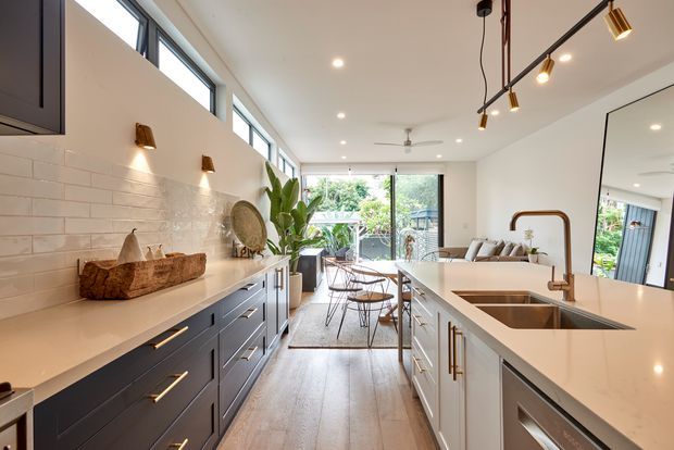 Bright open-plan contemporary kitchen with a large white island, integrated double sink and warm brass tapware, dark blue-grey cabinetry, white tiled splashback, and timber-look flooring. The kitchen opens to a dining and living area with abundant glazing, garden views, and a large wall mirror. Linear track lighting and brass accents add a refined modern character.
