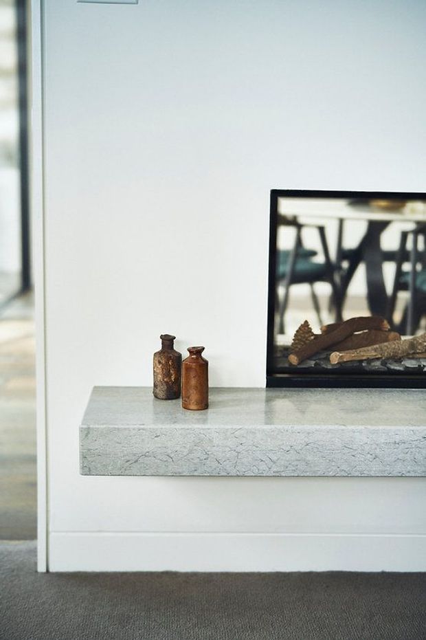 Close-up interior view of a minimalist fireplace installation. A thick pale grey stone hearth projects from a white wall, with a black-framed firebox and logs visible at right. Two small brown decorative vessels sit on the hearth, and a dark grey carpet covers the floor. A doorway or opening is visible at left.