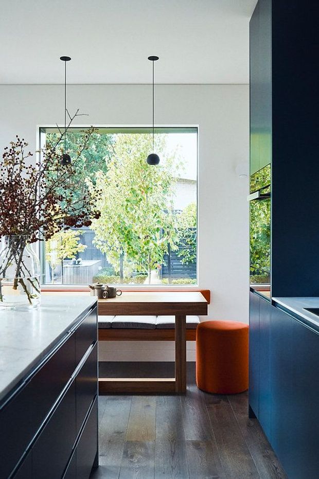 A bright contemporary kitchen with dark blue built-in cabinetry and a pale stone island. A large window overlooks greenery, while a timber breakfast bench with upholstered seating sits beneath it. Minimal black pendant lights, timber flooring, and a vivid orange cylindrical stool add contrast.