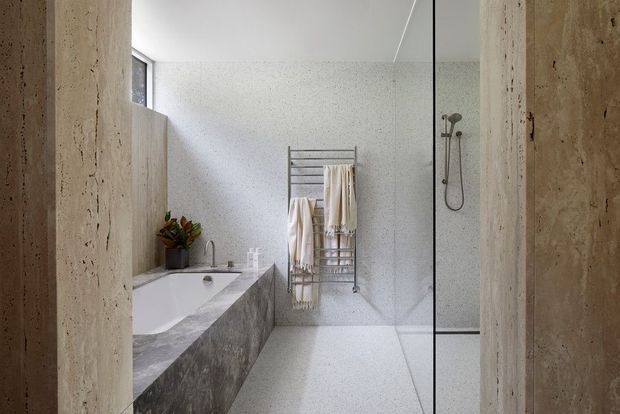 A refined contemporary bathroom viewed through a frameless glass shower enclosure. The space features a freestanding-style integrated bathtub with a dark grey stone-clad surround, pale terrazzo-like wall and floor finishes, a wall-mounted heated towel rail, and a minimalist exposed shower fitting. Large warm beige stone or travertine-clad vertical surfaces frame the room, creating a calm, luxurious atmosphere.