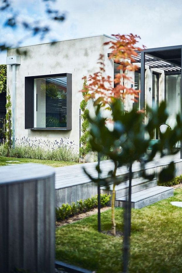 Exterior view of a contemporary single-storey home with a pale rendered facade, large dark-framed window, glazed entry or living area, and a covered outdoor deck. Layered timber decking, steps, lawn, and landscaped planting create a modern garden setting.