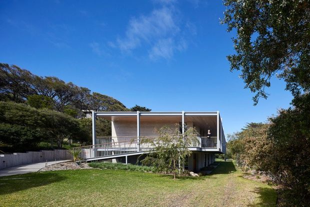 Exterior view of a contemporary elevated home set within a landscaped garden. The house features a lightweight rectangular form, broad open decks or verandahs with slim metal railings, large glazed openings, and a flat roof with timber-toned soffits. A ramp or gently sloping walkway leads toward the raised structure, surrounded by established trees and lawn under a clear blue sky.