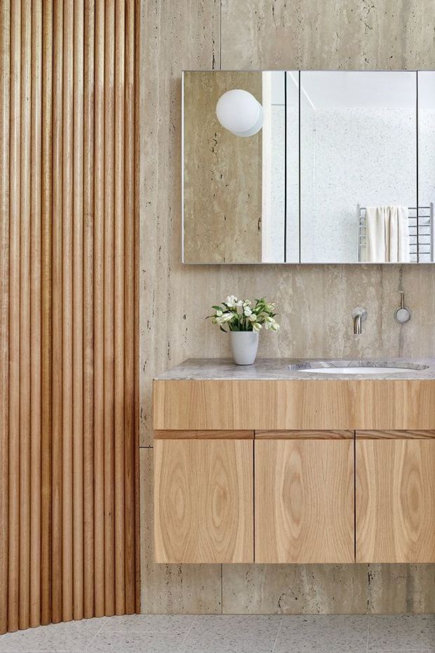 A refined contemporary bathroom vanity featuring a floating light timber cabinet with integrated basin and stone benchtop, set against a textured warm concrete or stone wall. A full-width mirrored cabinet, wall-mounted brushed metal tapware, round globe light, and vertical timber slat feature wall complete the calm, natural material palette.