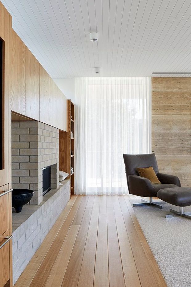 A bright, refined living room with a white-painted linear panel ceiling, warm timber wall panelling and flooring, and a large pale brick fireplace with integrated shelving. Sheer curtains cover a full-height window, while a dark leather lounge chair and matching ottoman sit on a light carpet beside the fireplace. The room has a calm mid-century-inspired character with natural materials and strong vertical and horizontal lines.