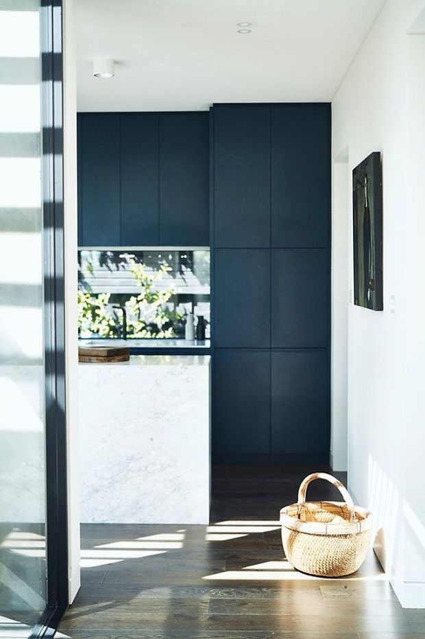 Bright contemporary interior view toward a compact kitchen, with tall matte dark navy cabinetry, a pale stone island or benchtop, dark timber flooring, white walls and ceiling, and strong sunlight cast through the glazing. A woven basket sits in the foreground, while an outdoor garden is visible beyond the kitchen window.