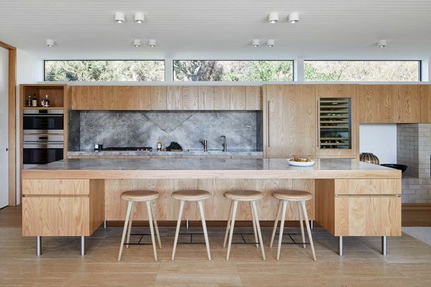 A bright contemporary kitchen with a long timber island and integrated seating for four. Full-height timber cabinetry spans the rear wall, with a dark stone-look splashback, integrated appliances, sink, open wine storage, and narrow clerestory windows beneath a ribbed ceiling. The kitchen opens toward a dining or living area at right, with a partial brick wall visible.