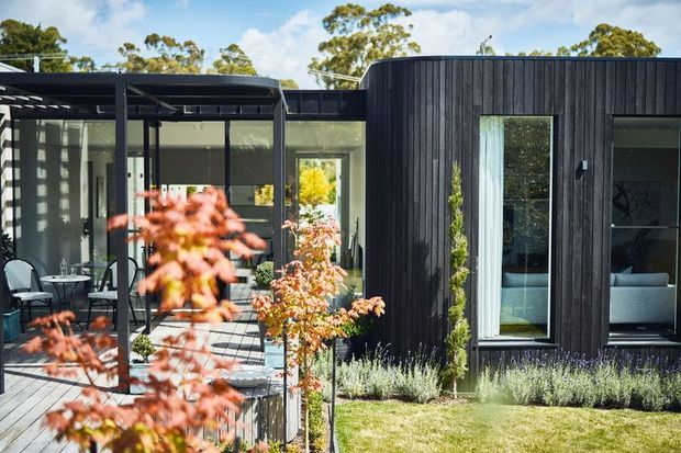 A contemporary single-storey home exterior with a dark vertically clad wing, expansive glazing, and a covered outdoor entertaining area. The courtyard-like garden includes structured planting and a lawn, while interior living spaces are visible through the windows.