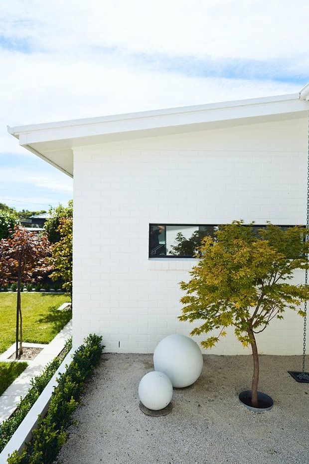 Bright exterior view of a minimalist white-painted brick building with a low-pitched roof and deep eaves. A narrow horizontal window is set into the wall, while a landscaped gravel courtyard contains clipped hedges, a Japanese maple, and spherical white ornaments. The image has a clean, modern residential character.