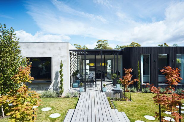 Single-storey contemporary residence viewed from the landscaped front garden, with a rectilinear composition of pale rendered walls and dark vertical cladding. A central glazed entry or outdoor transition space connects to a timber-look boardwalk, with neat lawns, stepping stones, and ornamental plantings. The home has a flat roofline and strong indoor-outdoor connection under a bright sky.