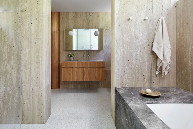 A refined contemporary bathroom with a freestanding bath in the foreground, extensive warm grey stone or concrete wall surfaces, and a dark stone bath surround. A timber floating vanity with integrated drawers, wall-mounted basin fittings, and a large mirrored cabinet are visible through the opening. The space has a minimalist, warm material palette with subtle vertical timber detailing and terrazzo-like flooring.