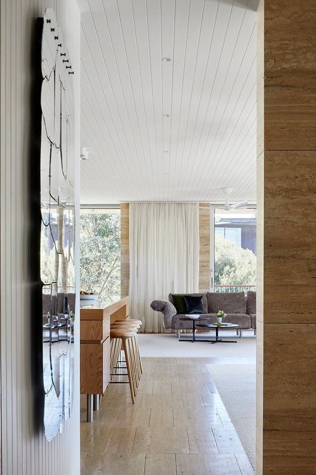 A bright open-plan interior viewed from a passage beside a kitchen or bar joinery area, leading into a relaxed living room. The space features white vertical wall and ceiling panelling, pale timber or stone-look flooring, warm wood cabinetry, a reflective dark backsplash or wall surface, a brown upholstered sofa, a black metal coffee table, and full-height windows with sheer curtains overlooking greenery. A textured warm stone-clad wall frames the right side.