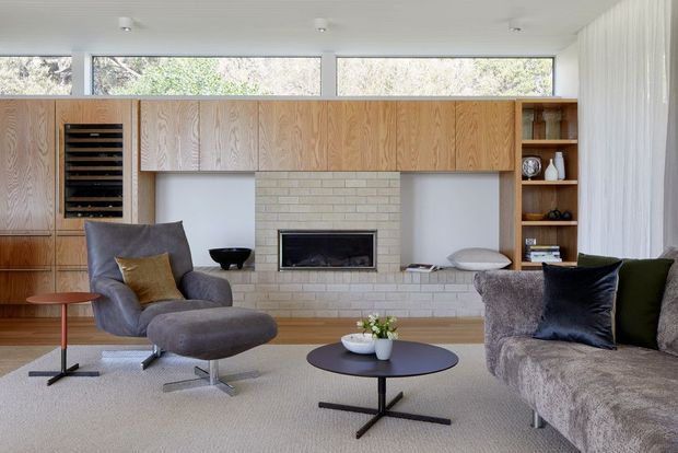 A bright mid-century-inspired living room with extensive warm timber built-in cabinetry, a central pale brick fireplace, high clerestory windows, and neutral upholstered lounge furniture arranged around low round coffee tables. The room has a calm, refined atmosphere and a compact, thoughtfully integrated media and storage wall.