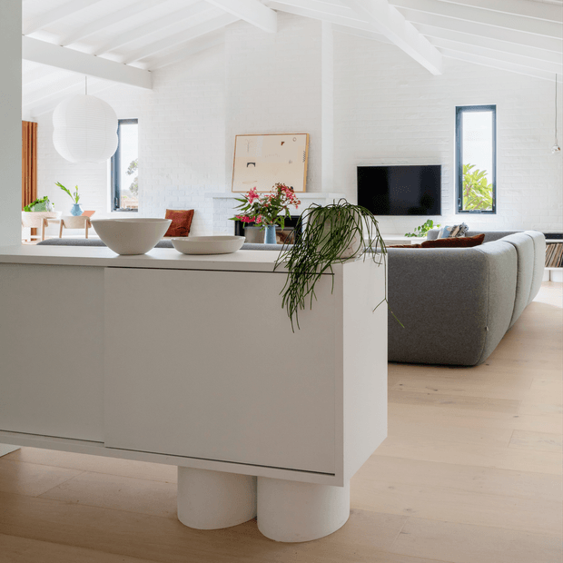 Bright open-plan interior with a white kitchen island in the foreground, pale timber flooring, white-painted brick walls and exposed white ceiling beams. The island has a smooth white benchtop, sculptural white pedestal supports, ceramic bowls and trailing greenery. A grey curved-back sofa, wall-mounted television, framed artwork, pendant light and large windows are visible in the living area beyond.