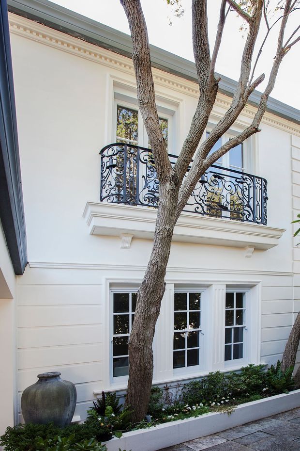 A refined white classical-style building facade viewed from a landscaped courtyard. The two-storey elevation features decorative cornice moulding, pilasters and trim, tall multi-pane windows, and an upper balcony with ornate black wrought-iron balustrading. Mature trees, a raised planted bed, a large urn, and stone paving occupy the foreground.