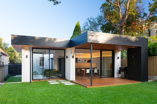 A contemporary single-storey rear extension or pavilion with a striking flat-roof form and dark horizontal cladding. Large floor-to-ceiling glazed openings connect the interior kitchen and dining area to a timber deck and manicured lawn. The structure combines white rendered walls, black/dark cladding, warm timber soffits and decking, and extensive black-framed glazing, with mature trees and neighbouring residential structures surrounding the site.