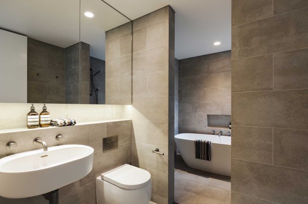 Contemporary bathroom featuring large-format taupe and grey stone-look wall and floor tiles, a floating white vanity basin, wall-hung toilet, freestanding white bathtub, recessed shelving, frameless mirror, and recessed ceiling lighting. The space has a clean, minimalist fit-out with a warm neutral palette and a partial visual connection between the vanity/toilet zone and bathtub area.