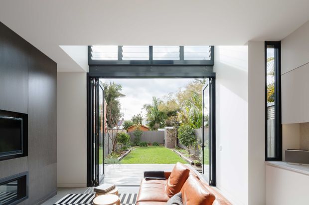 A bright contemporary living room opening directly onto a landscaped backyard through large black-framed folding or sliding glass doors. The room features a warm tan leather sofa, dark timber built-in cabinetry with a television and fireplace, pale walls and ceiling, light flooring, and a compact kitchen or joinery zone at right. High-level clerestory windows bring additional daylight into the space.