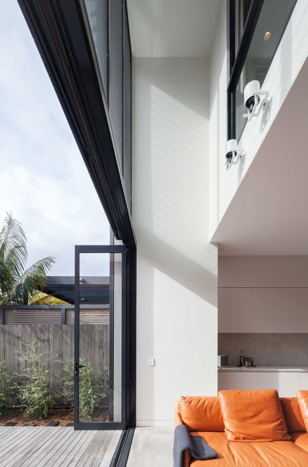 Bright contemporary interior viewed beside a large open black-framed glazed door. The image shows a double-height white-painted space with clean architectural lines, a partially visible minimalist kitchen, and a vivid orange leather sofa. Outside is a timber deck, privacy fencing, and tropical planting. Strong indoor-outdoor connection and abundant natural light dominate the composition.