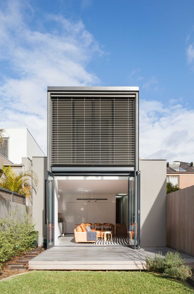 Exterior view of a narrow contemporary multi-level residence with a tall glazed opening, dark horizontal external louvres, and large folding or sliding glass doors opening onto a timber deck. A bright dining and living area is visible inside, with neighboring urban homes, boundary fencing, planting, and lawn surrounding the facade.