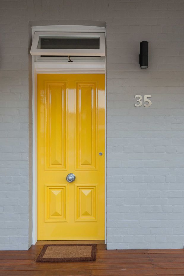 Close-up exterior view of a bright yellow, traditionally panelled front door with a silver round door knob, set in a light grey painted brick facade. A small transom window sits above the door, with a black cylindrical wall light and the house number 35 mounted beside it. The entrance opens onto a timber decking area with a rectangular doormat.