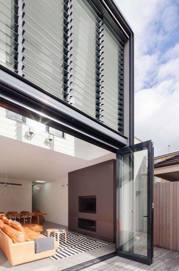 A bright contemporary living room opening onto a timber deck through a large black-framed glazed folding or pivot door. The room features a double-height glazed facade with external horizontal sun-shading fins, a warm orange leather sofa, timber dining furniture, a dark built-in media wall with fireplace, and a bold black-and-white striped rug. The composition also shows the adjoining timber-clad exterior and neighbouring roofline.