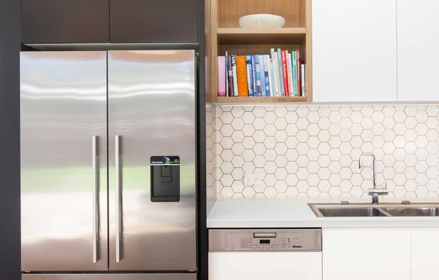 Close-up view of a contemporary kitchen featuring a large stainless-steel French-door refrigerator, dark upper cabinetry, white lower cabinetry, a white benchtop, and a white hexagonal tile splashback. Open timber shelving displays books and a ceramic bowl, while a stainless-steel sink and integrated dishwasher are visible.