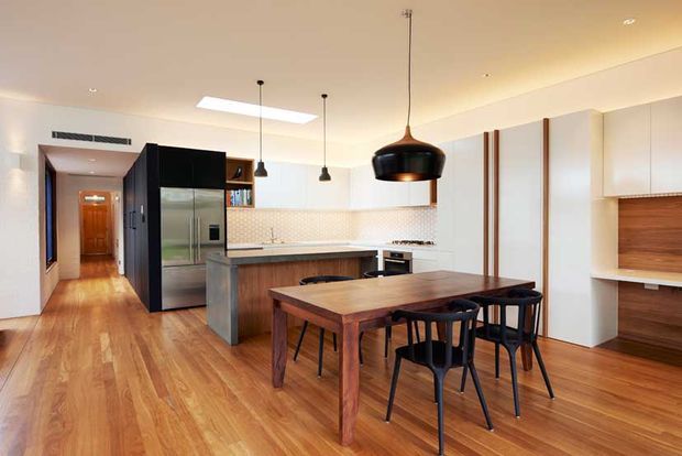 Open-plan kitchen and dining area with warm timber flooring, white full-height cabinetry, a central island, stainless-steel refrigerator, tiled backsplash, and a large timber dining table with black chairs. A black sculptural pendant hangs above the dining table, while additional pendant lights illuminate the kitchen. The space has a clean, warm contemporary feel with a skylight and hallway visible beyond.