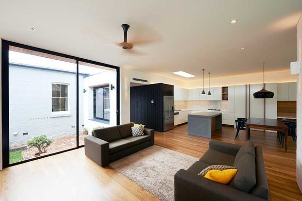 Bright open-plan living, dining and kitchen area with large black-framed glazing opening onto a small courtyard. The room features warm timber flooring, dark charcoal sofas, a neutral rug, a compact grey kitchen island, white flat-panel cabinetry, dark wood-toned joinery, pendant lights and a ceiling fan.