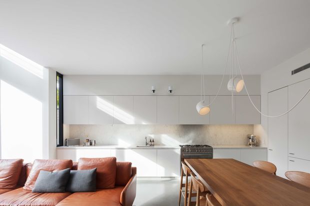 Bright open-plan kitchen and living area with a minimalist white kitchen, integrated cabinetry, stone-look splashback, freestanding range cooker, timber dining table with chairs, and a contemporary leather sofa. Large windows and sculptural pendant lights create an airy, sunlit interior.